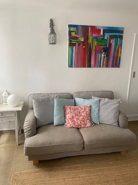 A cozy living area is presented with a light gray sofa adorned with colorful cushions, including a coral-patterned accent cushion. Above, an abstract painting adds color to the white wall, while a small side table with decorative items complements the space.