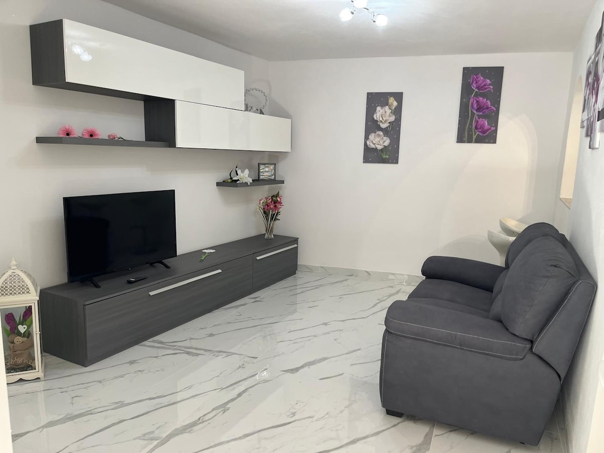 A modern living area features a sleek gray couch and a low profile entertainment unit. A wall-mounted television is positioned opposite the seating. Floral artwork adorns the walls, and a smooth, light-colored floor enhances the space's brightness.
