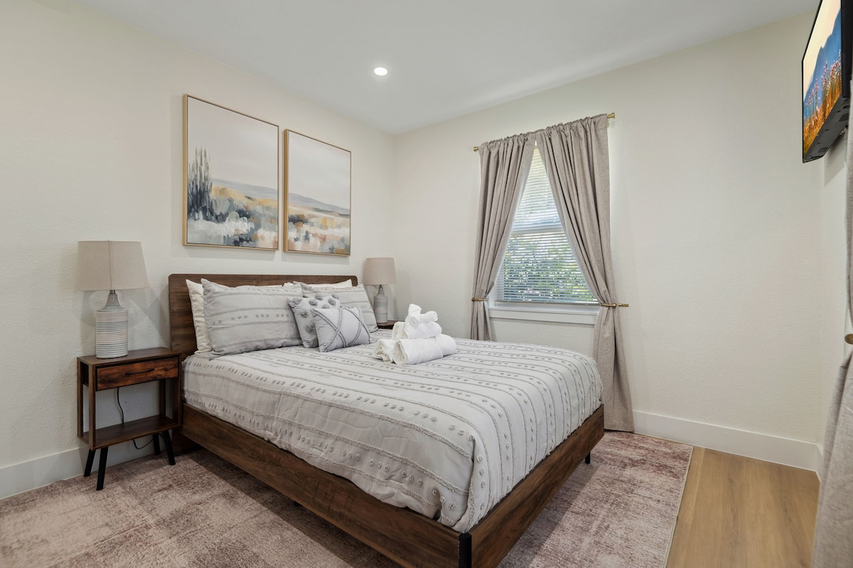 A comfortable sleeping area features a spacious bed dressed in soft linens. Two elegant wall art pieces above the bed complement the neutral tones. A window with light curtains allows natural light to enter, while two bedside lamps add functional illumination.