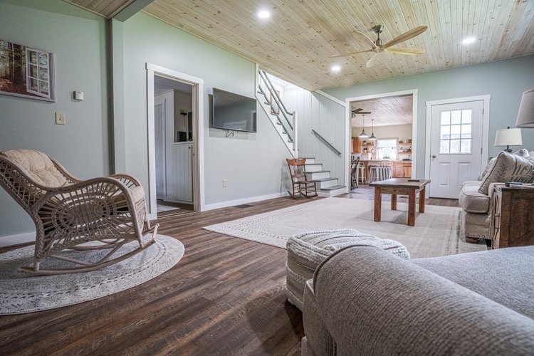 A spacious living area is highlighted by a light wood ceiling and natural light from multiple windows. Comfortable seating options include a textured armchair and a plush couch. A wooden coffee table sits centered on a soft area rug, while stairs lead to the upper level.