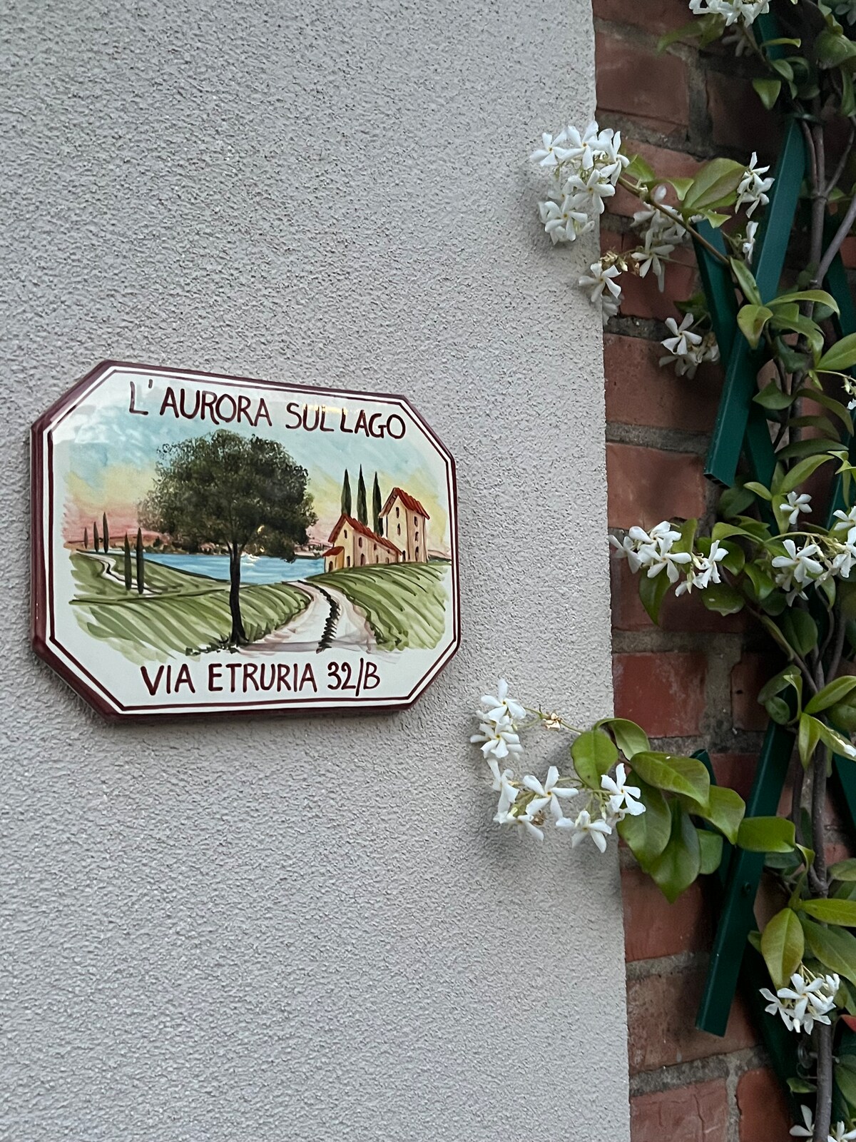 A painted ceramic house sign displays the name 'L'Aurora Sul Lago' along with the address 'Via Etruria 32/B'. Surrounding the sign, white flowers and green vines add a natural touch against the texture of the wall.