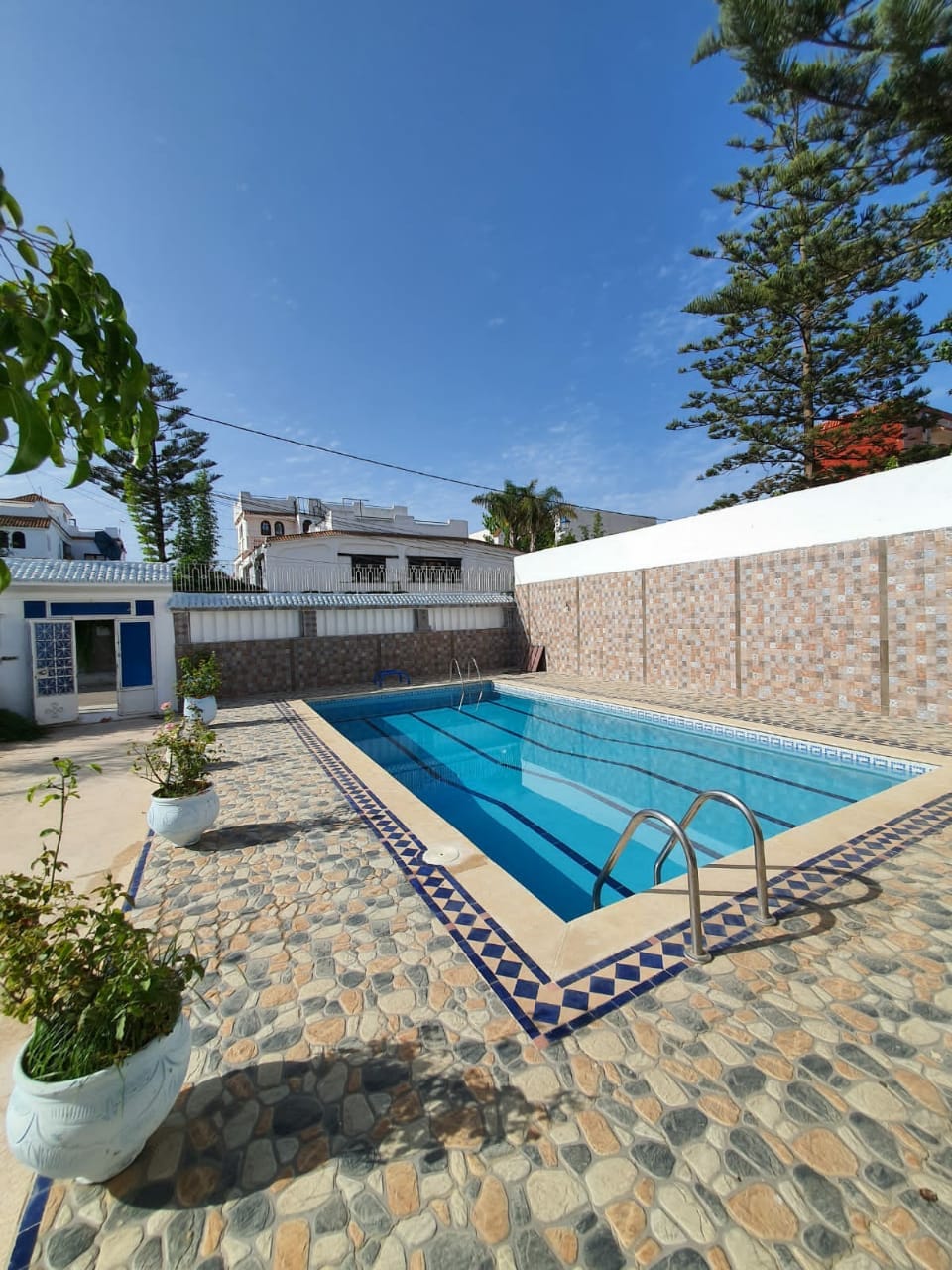 A large swimming pool is set within a tiled area, framed by planters with greenery. The surrounding walls feature textured stonework, with a clear blue sky above and trees in the background, providing a serene outdoor environment.