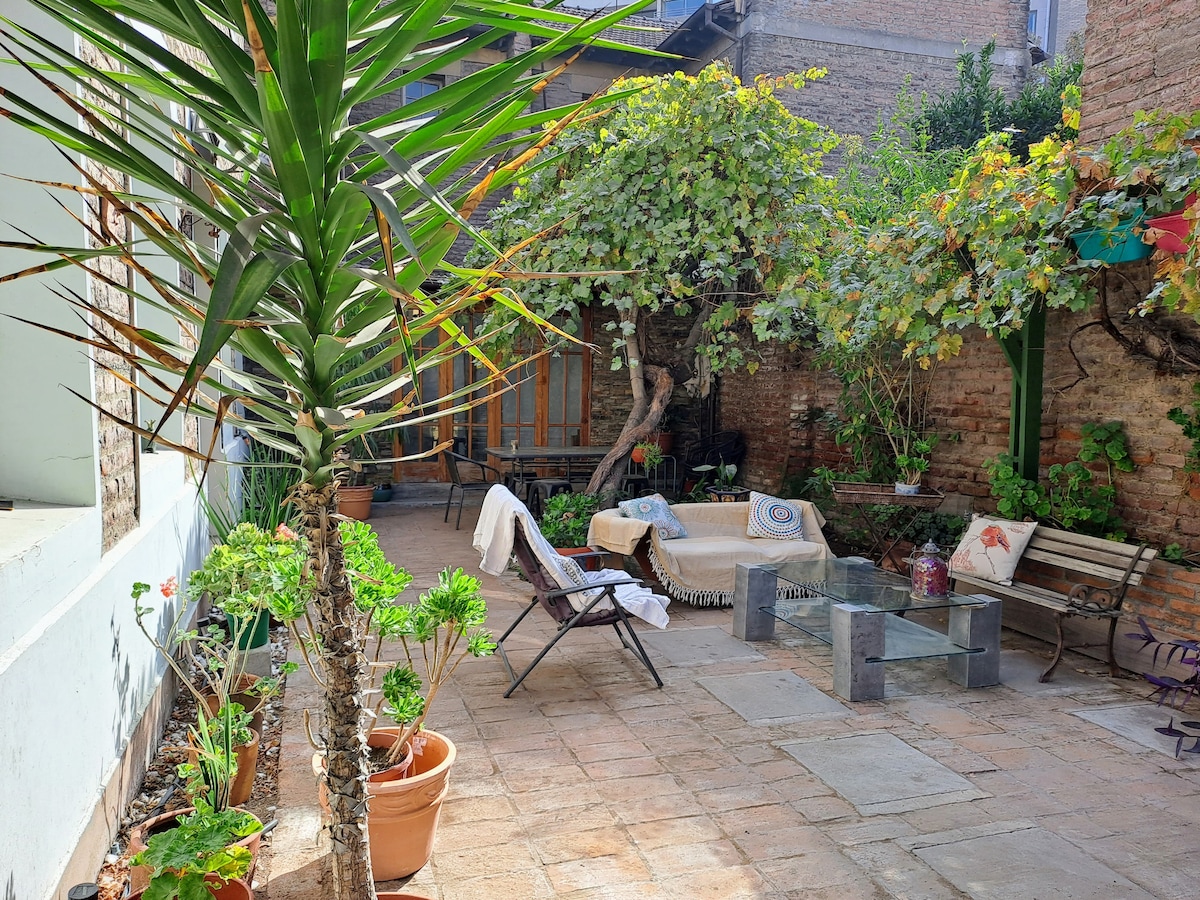 A shared patio area is shown, featuring stone flooring and a variety of plants. Comfortable seating arrangements include wooden chairs and a low table, surrounded by climbing vines. A cozy throw and decorative pillows are placed on the furniture, enhancing the inviting environment.