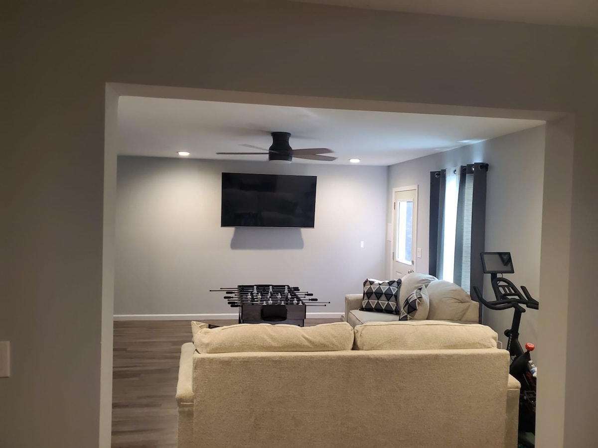 A spacious living area is presented, featuring a light-colored couch facing a mounted television. A foosball table is positioned nearby, and a ceiling fan gently circulates air. Natural light enters through a window dressed with dark curtains.