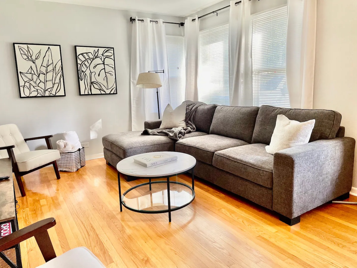 A cozy living area features a spacious gray sectional sofa, accompanied by a round coffee table. Two large windows with sheer white curtains allow natural light to brighten the space, while two framed art pieces adorn the white walls.