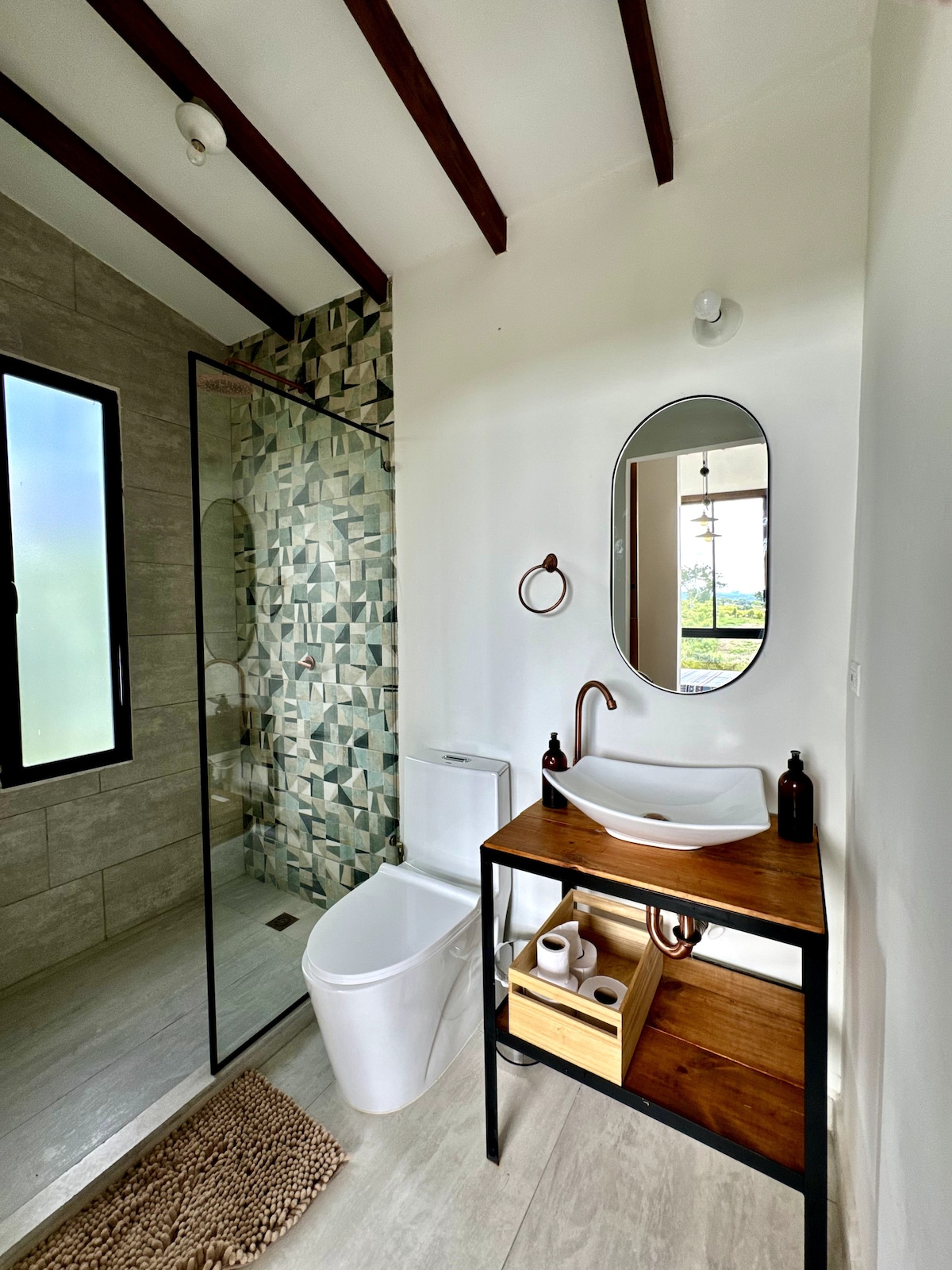 A modern bathroom is featured with a standing shower enclosed in glass. A stylish wooden vanity with a round mirror and a spacious sink is visible, alongside storage for toiletries. Natural light filters through a window, highlighting the textured wall tiles.