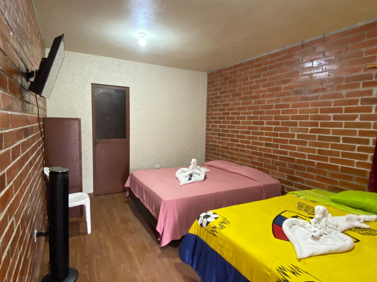 A cozy sleeping area features two beds, one with a pink cover and the other with vibrant yellow and blue bedding. Decorative towel arrangements add a unique touch. A small television is mounted on the wall, and a fan stands beside a white chair on the wooden floor.