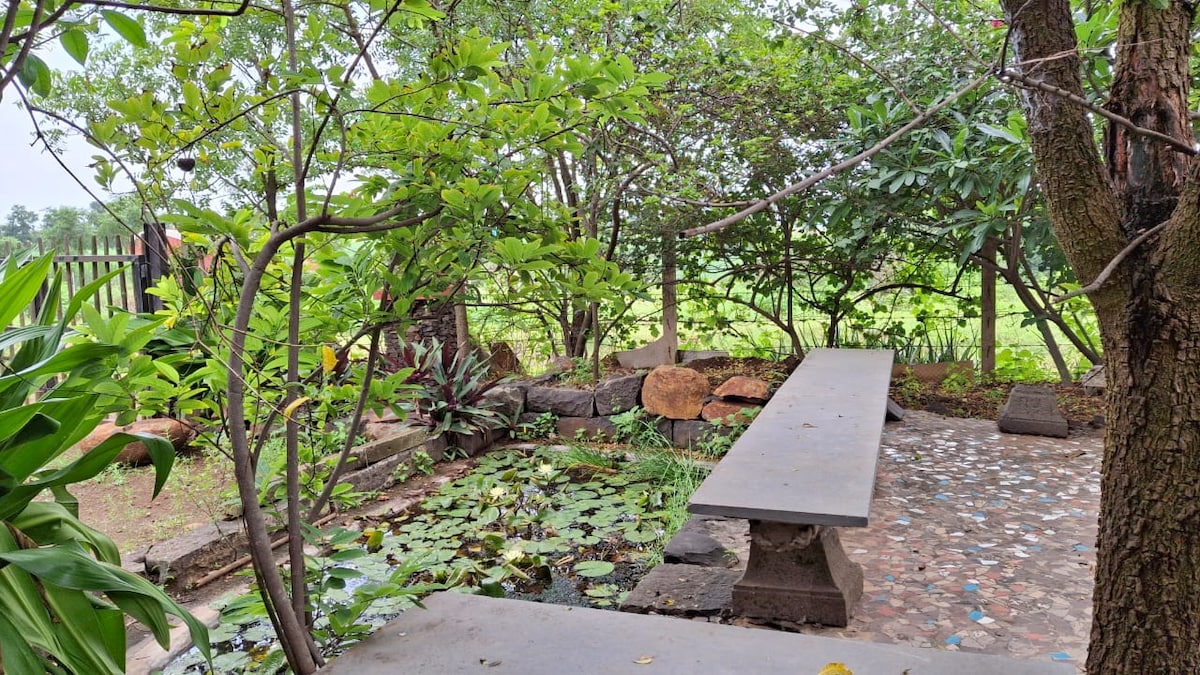 A serene garden area features a long stone bench amidst lush greenery and various plants. A small pond with lily pads adds a tranquil element, while surrounding trees provide natural shade. The inviting space is perfect for relaxation and enjoying the surrounding landscape.