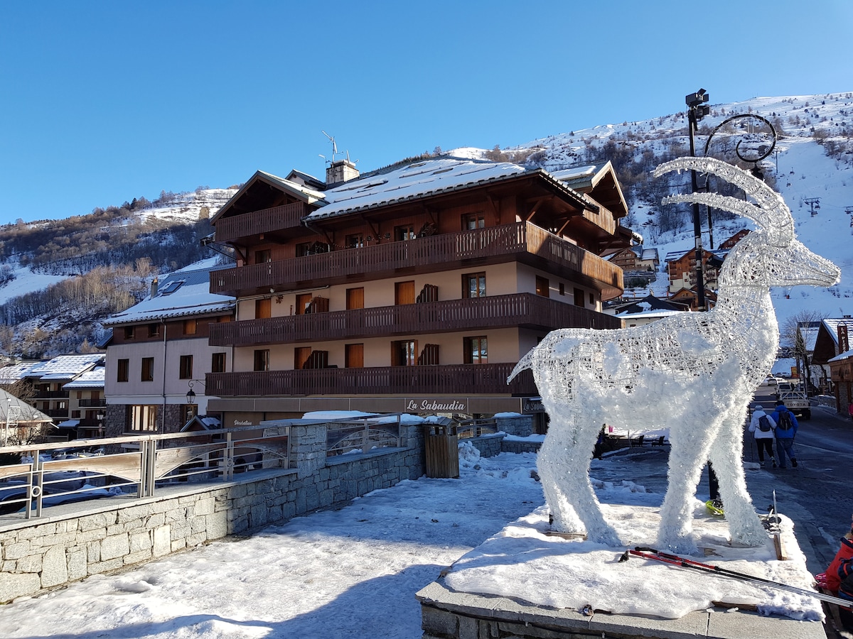 A multi-story building is depicted, showcasing traditional alpine architecture with wooden balconies and a snow-covered roof. A decorative reindeer sculpture is positioned in the foreground, adding a festive touch to the winter scene. The surrounding landscape features snow-capped mountains.