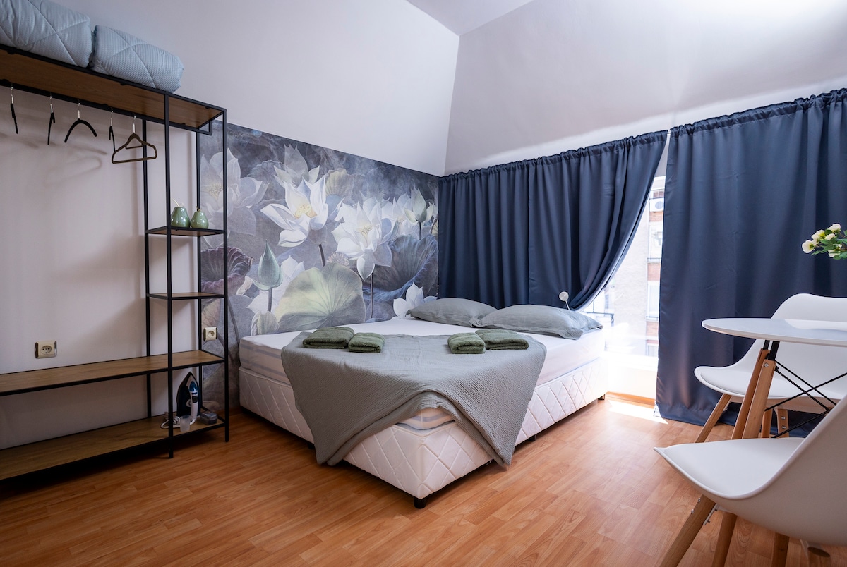 A compact studio features a spacious bed with soft gray linens and dark green towels neatly arranged. A floral mural adorns the wall, complemented by dark curtains framing the window. A small dining area with white chairs is positioned beside a shelf, adding functionality to the space.