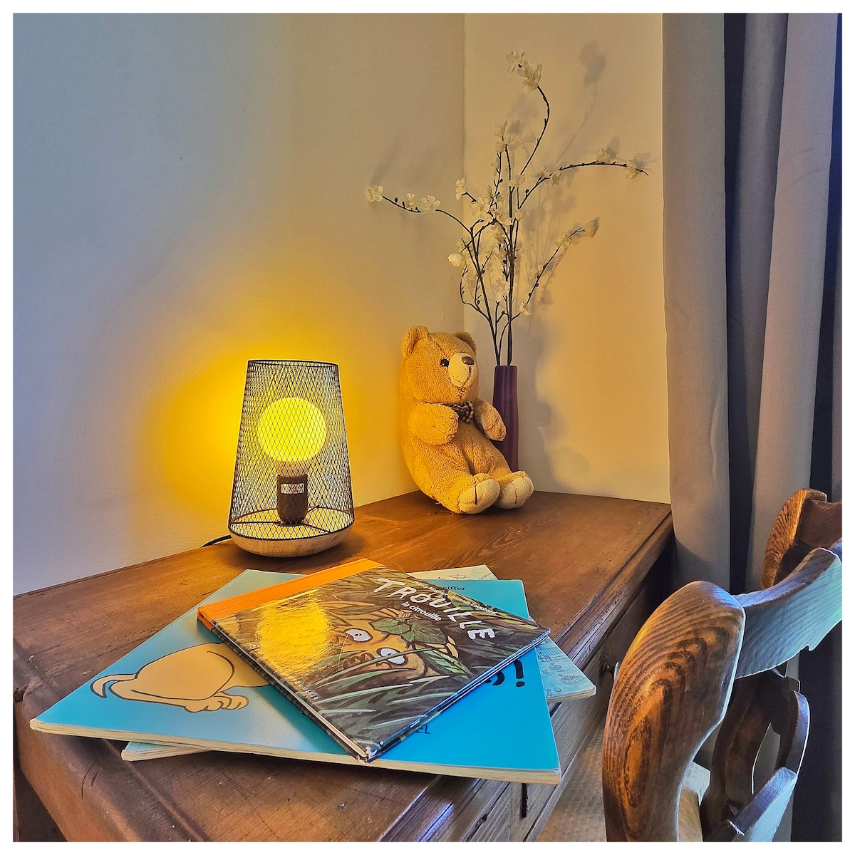 A wooden table is adorned with a decorative lamp casting a warm glow. Books are stacked on the surface, and a teddy bear sits nearby alongside a small arrangement of artificial flowers, contributing a homely touch to the space.