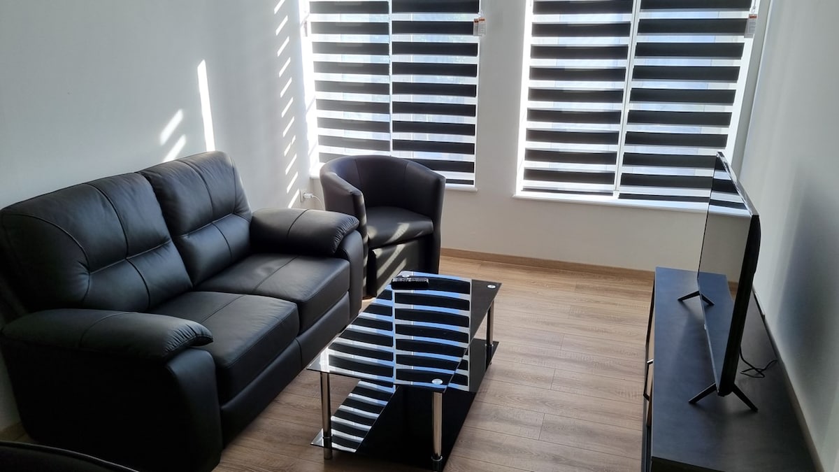 A living room is presented with a black leather sofa and two matching armchairs positioned around a glass coffee table. Natural light filters through the neatly arranged black and white striped shades on the large windows, complementing the warm wooden flooring.