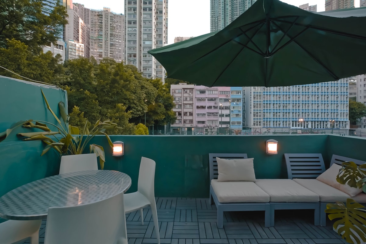 A private rooftop area features a modern lounge with a sectional sofa and a round dining table. A large umbrella provides shade, while urban views of surrounding buildings and greenery are visible in the background. Soft lighting is provided by wall-mounted fixtures.