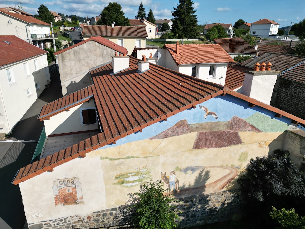 Aerial view of a village featuring a mural on a prominent wall. The mural depicts a landscape with mountains and figures engaged in agricultural activities. Red-tiled roofs of surrounding buildings are visible, along with trees and greenery nearby.