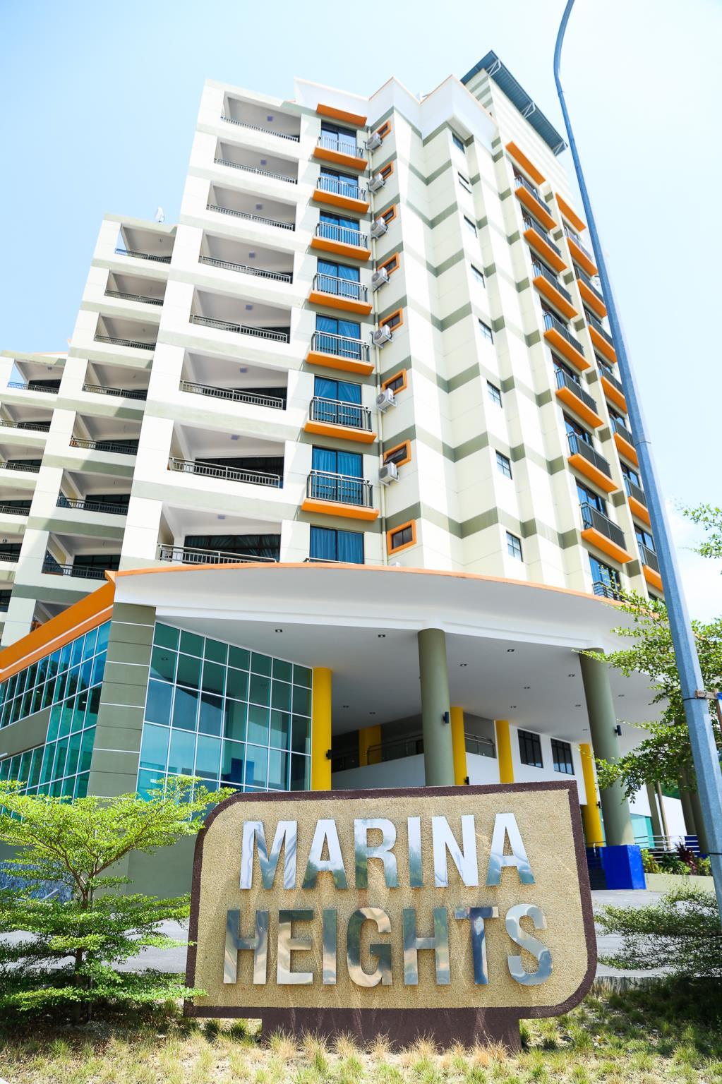 A modern building named Marina Heights is depicted, featuring multiple stories with large windows. The entrance is accentuated by a prominent sign at the forefront, accompanied by green landscaping. The façade showcases a combination of light and vibrant colors, enhancing its contemporary design.