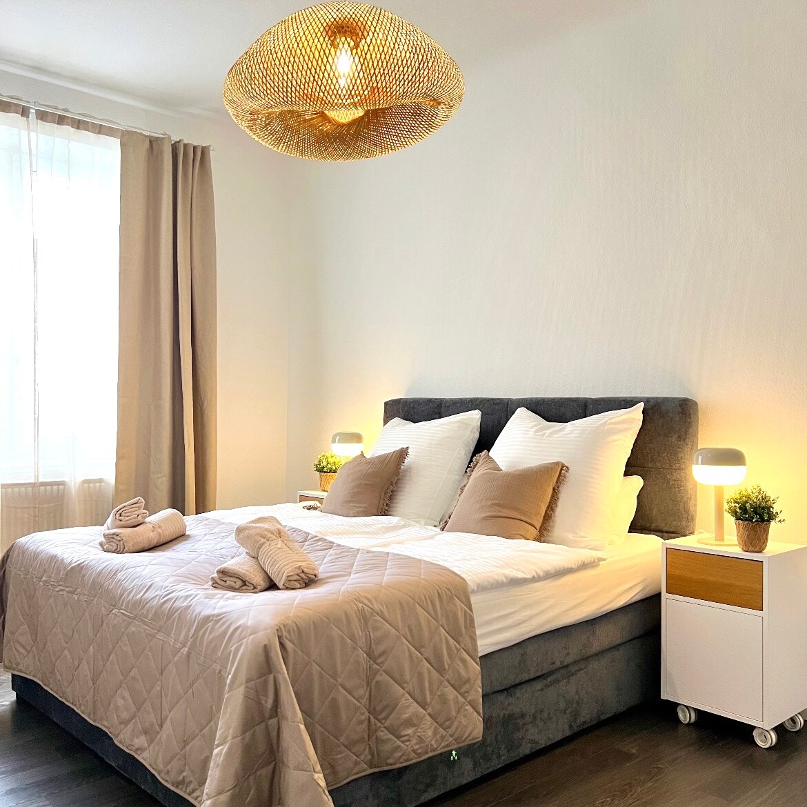 A well-appointed bedroom features a spacious bed with crisp linens and decorative pillows. Soft lighting is provided by two bedside lamps, and a stylish woven light fixture hangs overhead. Natural light filters through sheer curtains, creating a serene ambiance.