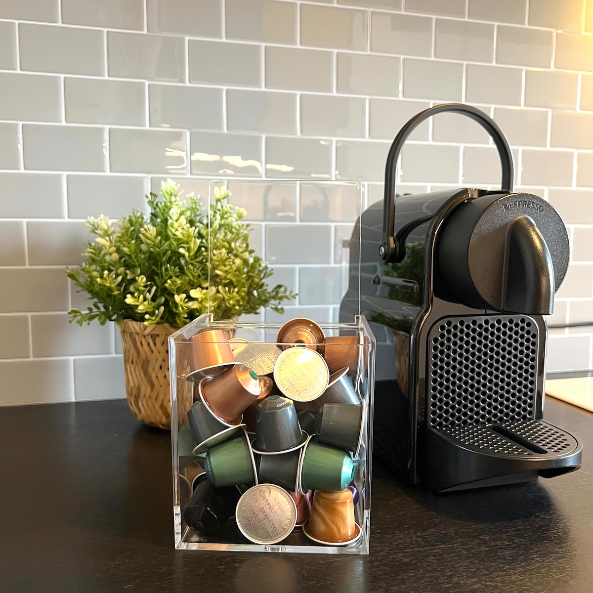 A Nespresso coffee machine is displayed alongside a transparent container filled with various coffee capsules in different colors. A small green potted plant adds a touch of greenery against a backdrop of light gray tiled walls.