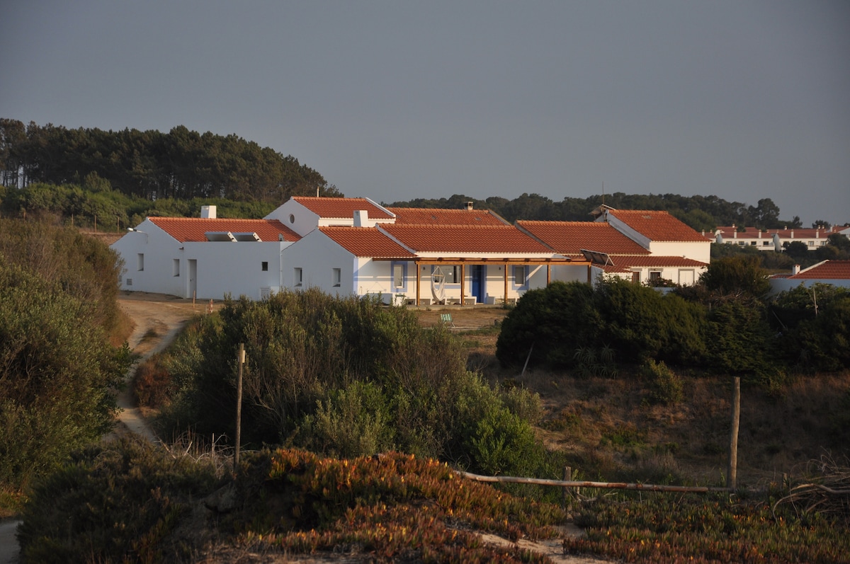 A charming white structure featuring red-tiled roofs is nestled among lush greenery. The building comprises multiple sections, with a pathway leading to a welcoming entrance. The surrounding landscape includes low shrubs and a glimpse of distant trees, offering a serene natural setting.