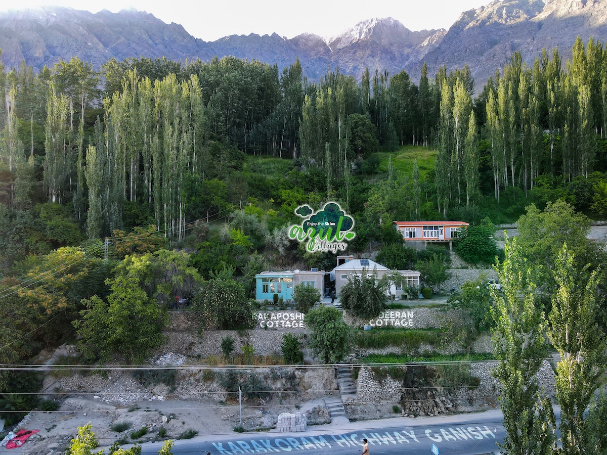 Two cottages are positioned amid lush greenery, showcasing a harmonious blend with the surrounding landscape. The cottages feature charming exteriors, and panoramic mountain views create a serene backdrop. A winding road can be seen in the foreground, providing access to the cottages.