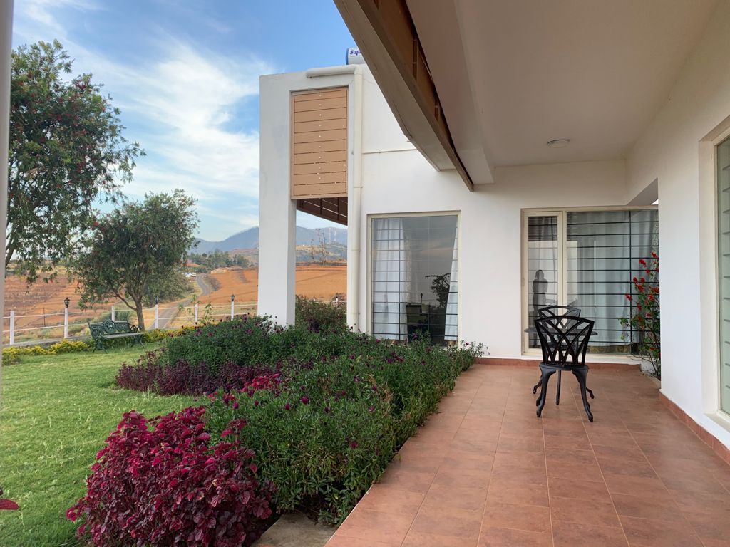An inviting outdoor sitting area is featured on a covered porch, overlooking a landscaped garden with colorful floral borders. A single black chair is positioned on the tiled surface, with large windows allowing natural light into the nearby interior spaces and a scenic hillside visible in the background.