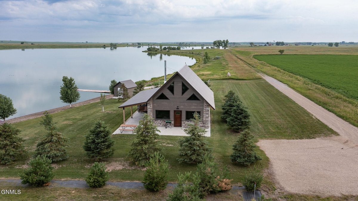 A spacious cabin is positioned near a calm body of water, surrounded by grassy fields and trees. The structure features a triangular roof and outdoor seating arrangements on a patio. A winding gravel path leads up to the property, enhancing the sense of seclusion.