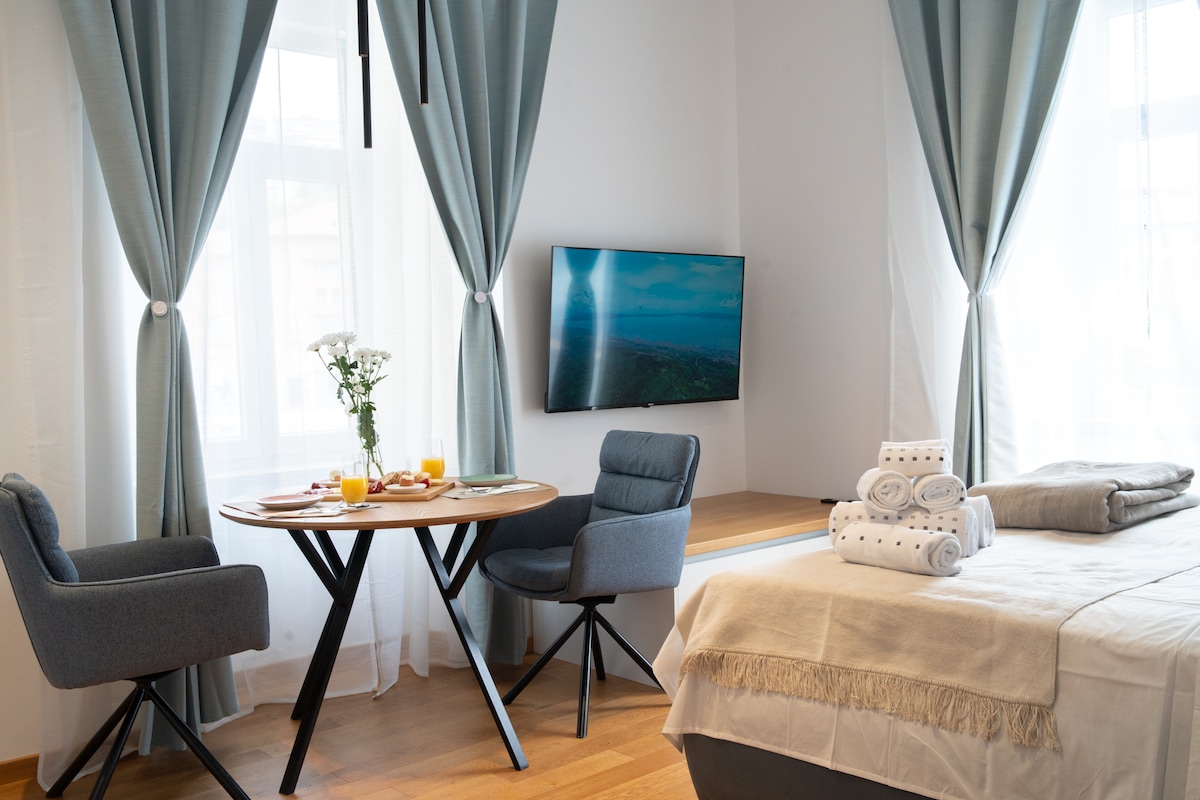 A cozy living space features a spacious bed with neutral linens. A wooden table is paired with two upholstered chairs, with a small vase of flowers at the center. Natural light is filtered through floor-to-ceiling curtains, and a mounted television adds convenience.
