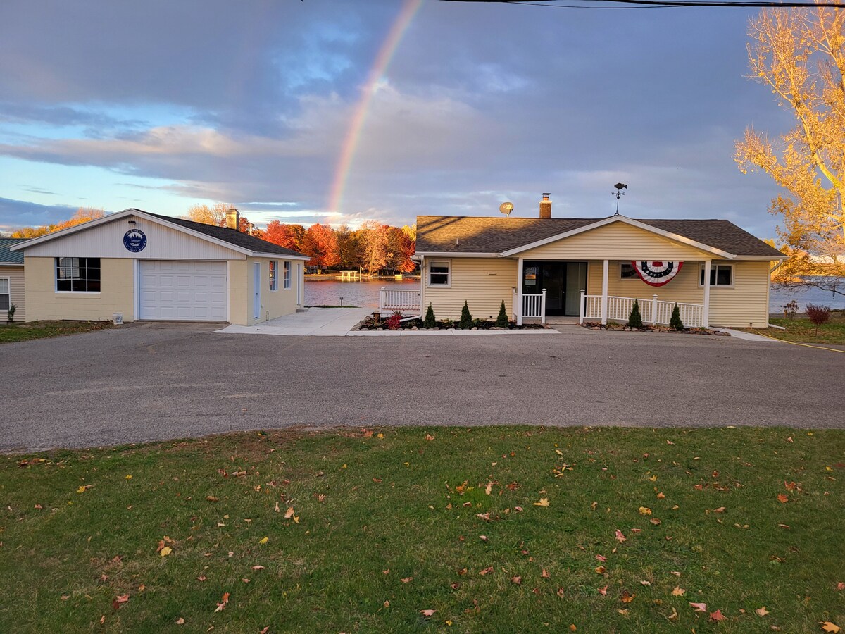A modern cottage is seen alongside a paved driveway, with a large garage structure visible on the left. A rainbow arcs over the home, while colorful autumn trees line the background. The property offers a welcoming façade with a front porch adorned with decorations.