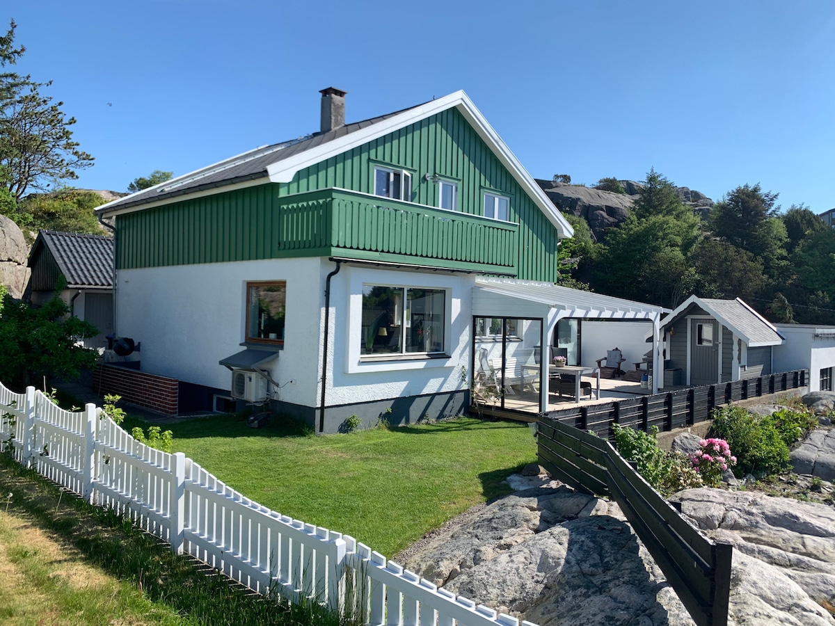 The exterior view showcases a three-story house with a green upper section and white lower walls. A spacious terrace is visible, framed by a white fence and surrounded by a manicured lawn. Natural rocky terrain and greenery enhance the setting, indicative of a peaceful outdoor atmosphere.