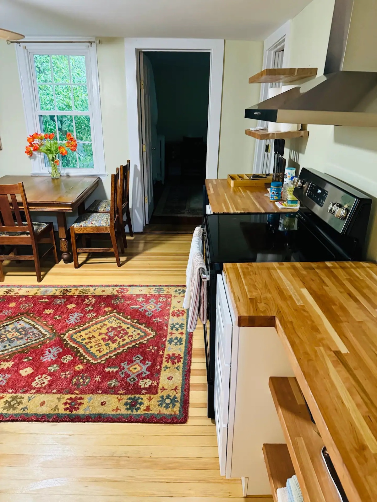 A modern eat-in kitchen is featured, showcasing wood countertops, a black stove, and a wooden table surrounded by chairs. A colorful area rug adds warmth, while a vase of flowers brightens the space. Natural light filters in through the windows, enhancing the inviting ambiance.