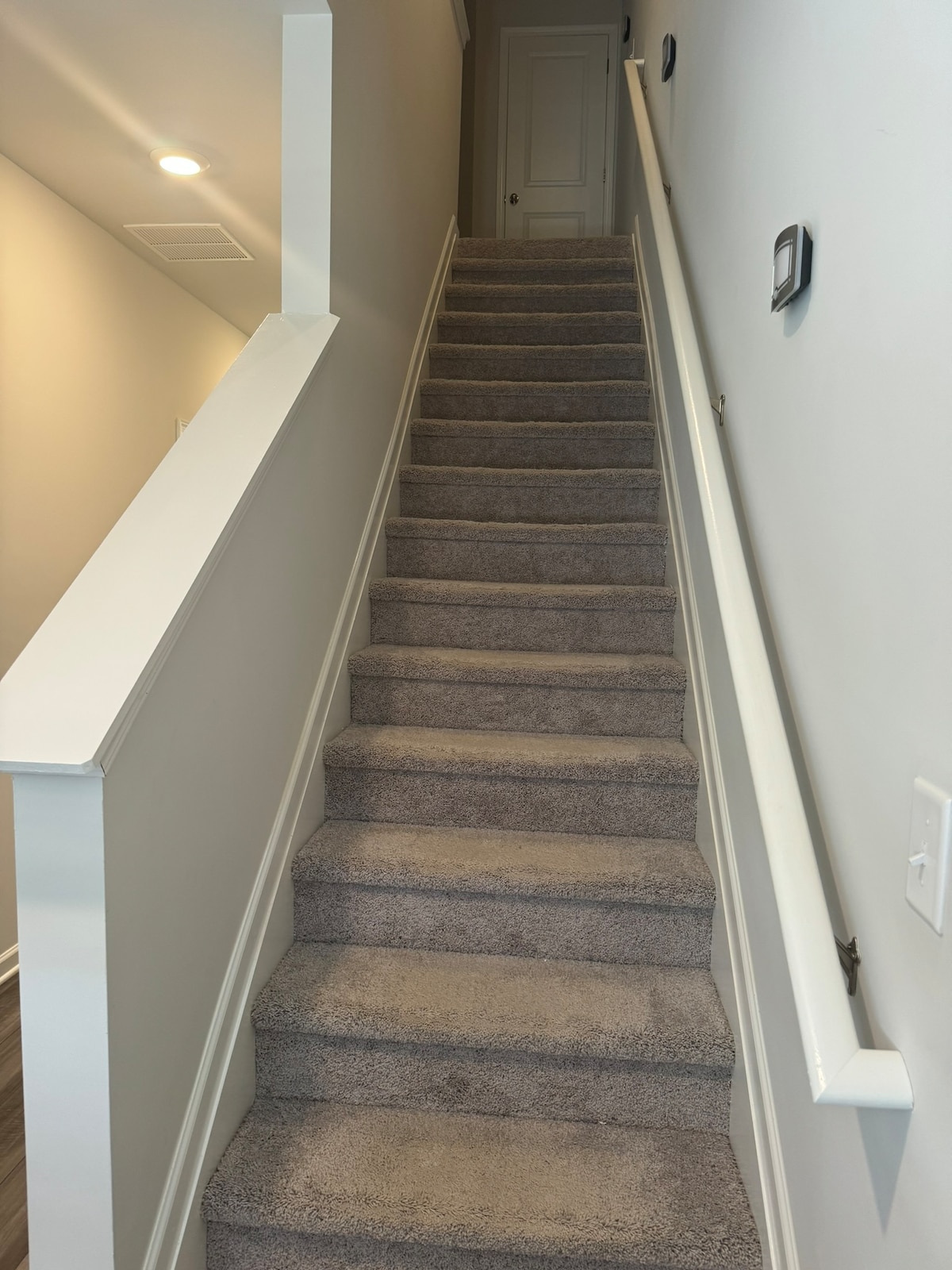 A carpeted staircase with neutral-colored walls leads to the upper level of the house. Handrails are present on both sides for safety. Soft lighting is provided by ceiling fixtures, illuminating the steps and enhancing visibility.