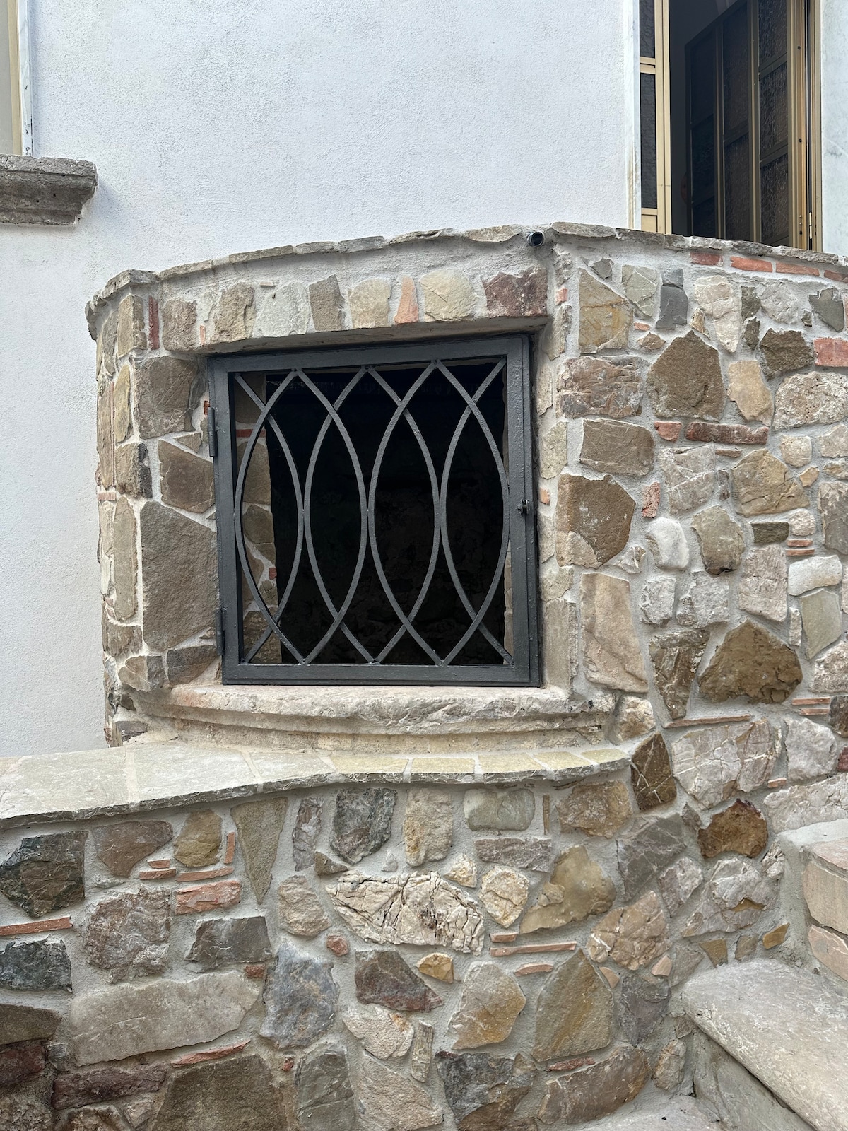 A stone wall showcases a rounded window framed with elegant ironwork. The natural stone surface features various shades and textures, enhancing the building's historic character. Stairs lead up to the entrance, indicating a welcoming approach to the property.