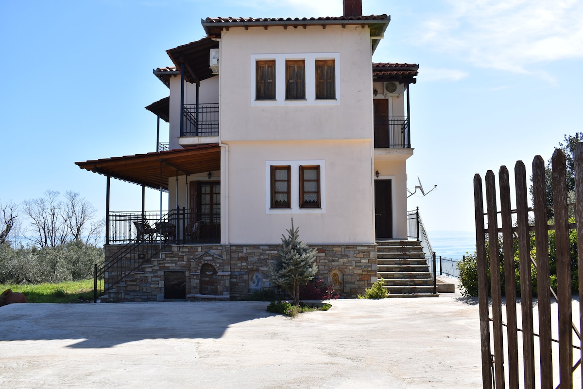 A two-story villa is presented, featuring a light-colored façade with multiple windows and a spacious entrance. A covered porch extends across the front, while steps lead to the main door. The surrounding area includes a gravel driveway and hints of greenery in the background.