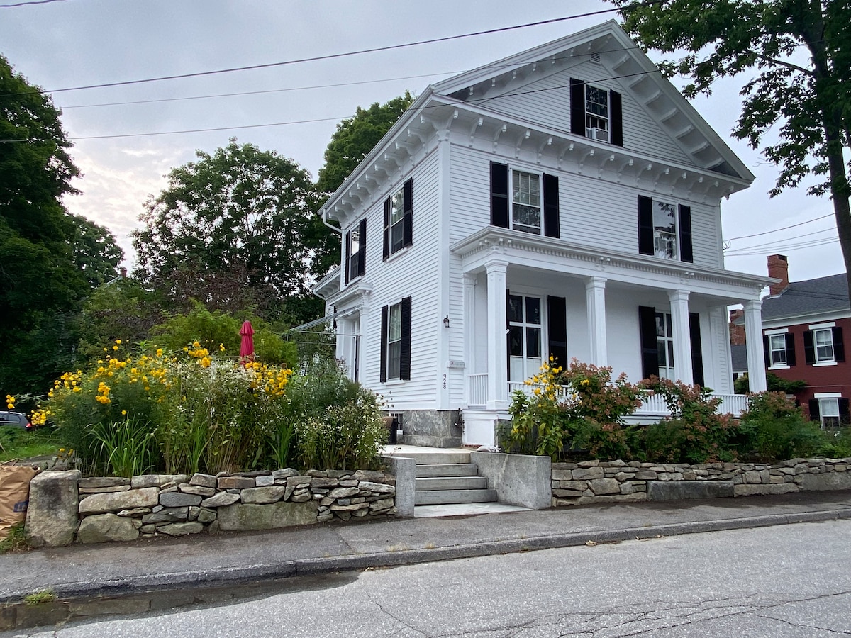 A two-story Greek Revival house is featured, showcasing white siding and prominent black shutters. The front porch is adorned with columns, and bright yellow flowers bloom in the garden beds, enhancing the home's curb appeal. A stone wall defines the landscaping along the sidewalk.