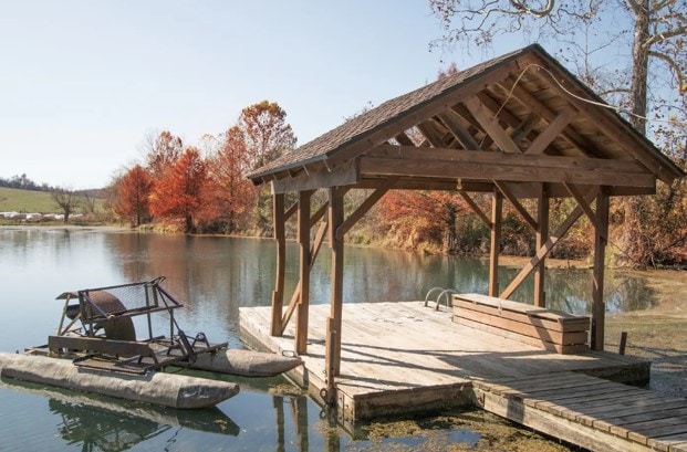 A wooden dock extends into calm waters, featuring a covered pavilion with a sloped roof. A fishing or paddle board rests on a flat surface next to the dock, with autumn foliage reflecting in the water, creating a serene landscape.