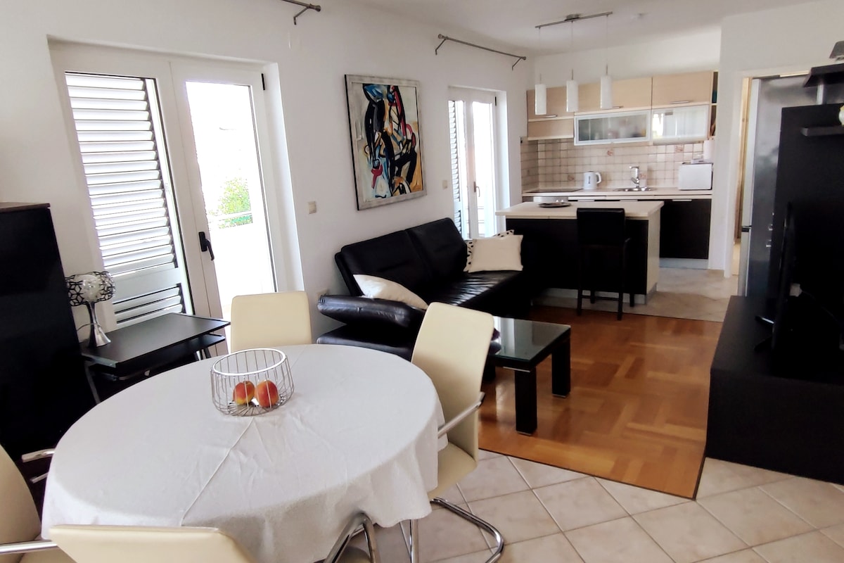 An open-plan living area features a dining table with a white tablecloth and a bowl of fruit. Adjacent, a sleek black sofa sits beside a modern kitchen equipped with light cabinetry and stainless steel appliances. Natural light streams through the sliding doors, enhancing the airy feel.