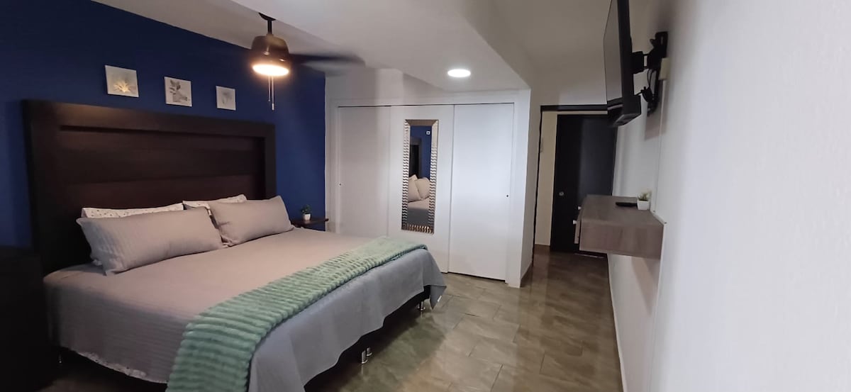 A spacious bedroom features a large, dark wooden bed with gray bedding and a light green throw. A mounted television is positioned on the wall opposite the bed. Light-colored tiles cover the floor, and a closet with sliding doors is visible in the background.