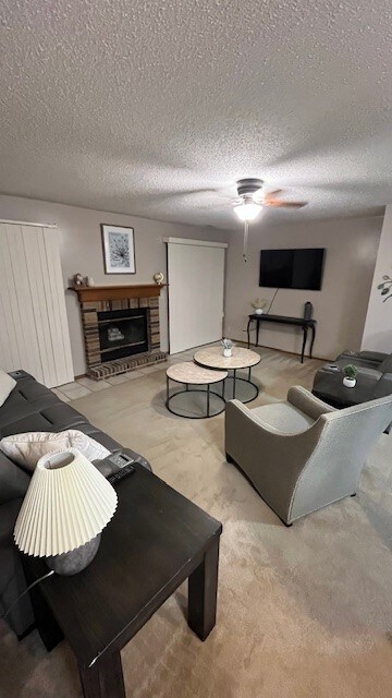 A cozy living area is furnished with a sectional sofa, a pair of accent chairs, and a coffee table. A fireplace is framed in brick, adding warmth. Natural light enters through sliding doors, complementing the neutral wall colour and soft carpeting.