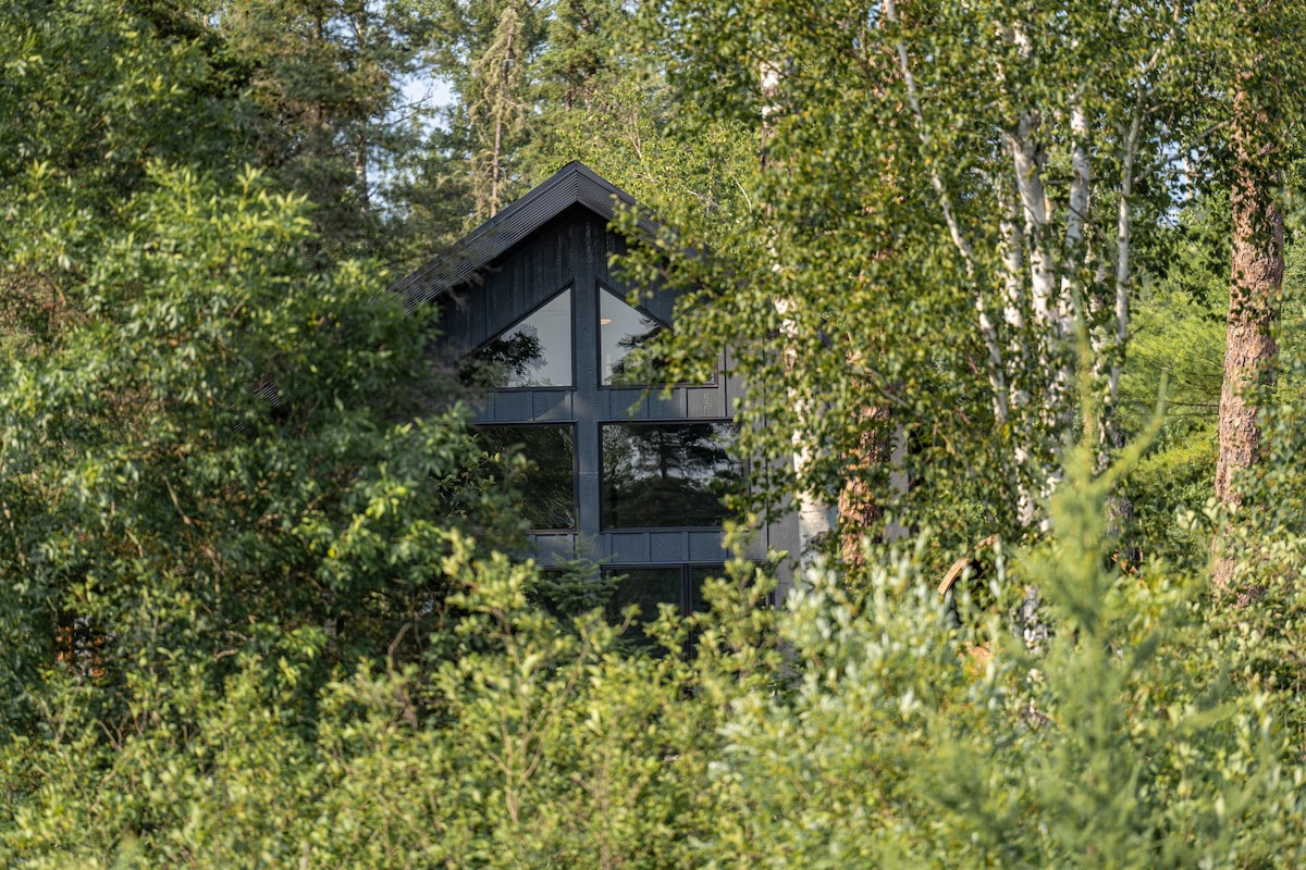 A modern cabin is nestled among lush greenery, featuring large glass windows that reflect the surrounding trees. The architectural design combines dark wood with a sloped roof, blending harmoniously into the forested landscape.