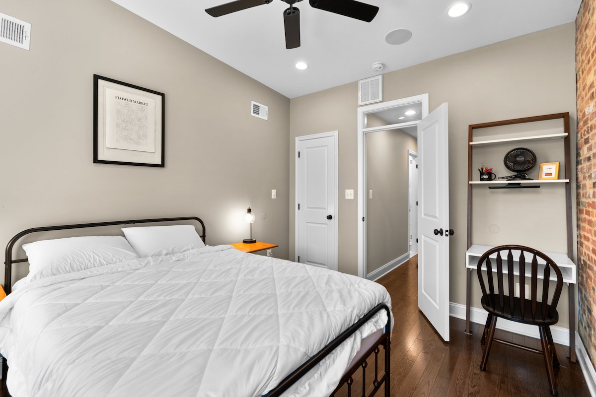 A well-appointed bedroom features a spacious bed with a minimalist white comforter. A wooden chair and desk are positioned near the wall, while a nightstand with a lamp complements the room’s neutral tones, including a highlighted exposed brick accent.