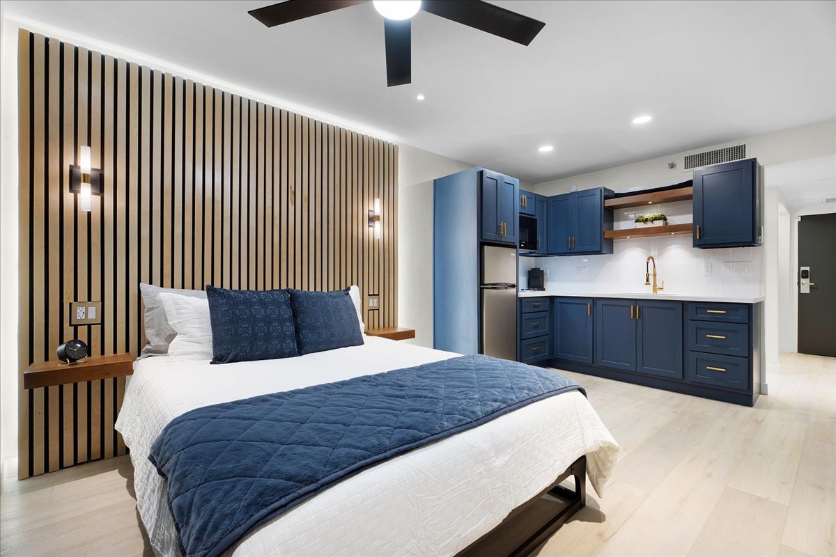 The studio features a spacious bed with crisp linens, complemented by a stylish headboard and accent wall of vertical wood panels. A modern kitchen area with blue cabinetry, stainless steel appliances, and a white countertop stands in the background, providing a sleek, functional design.