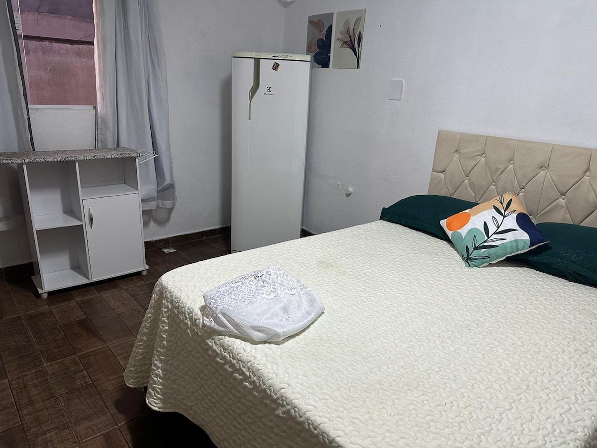A compact room features a light-toned bedspread on a spacious bed, flanked by green pillows. A small refrigerator stands next to a minimalist white table, while a sheer curtain allows soft light to enter from a nearby window.