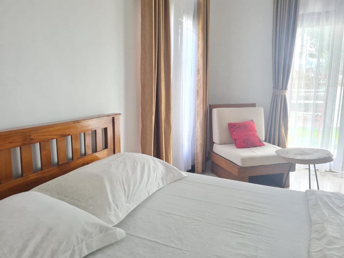A bright and airy bedroom features a spacious bed with crisp white linens. A cozy seating area includes a comfortable chair and a small table. Large windows are adorned with soft curtains, allowing natural light to fill the room and providing views of the outside garden.