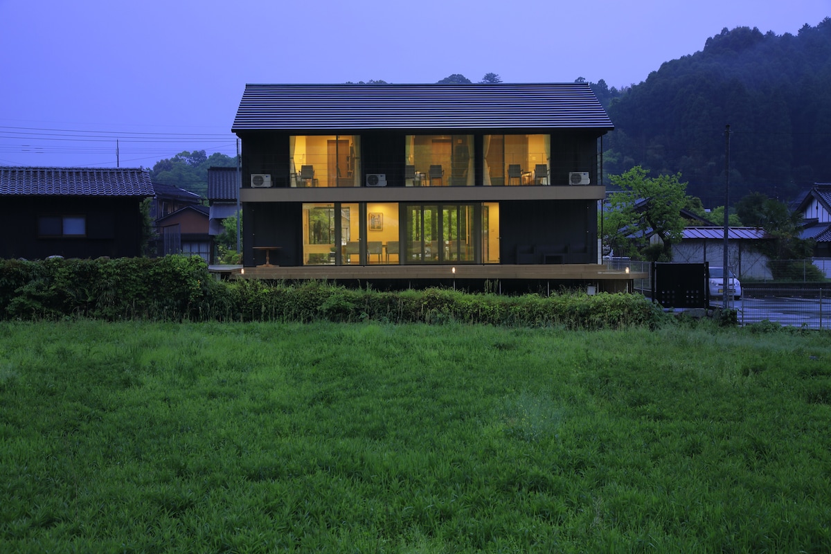 A modern two-story building is shown at dusk, with warm light emanating from large windows. The structure is surrounded by a lush green field, and gentle hills rise in the background, creating a peaceful rural setting.