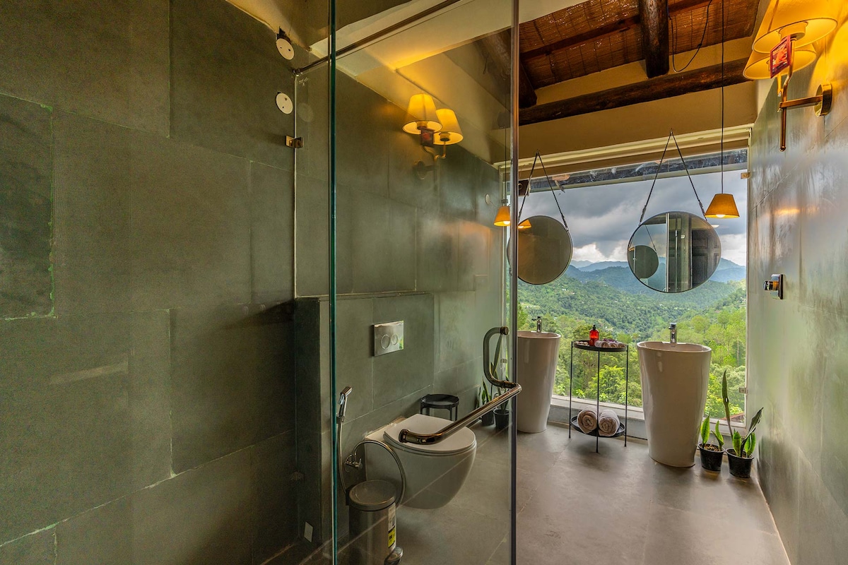 A spacious ensuite bathroom features large glass windows that offer scenic views of the mountains. Two freestanding bathtubs are illuminated by pendant lighting, with a modern vanity and clean lines throughout. The stone-clad walls create a serene and contemporary ambiance.