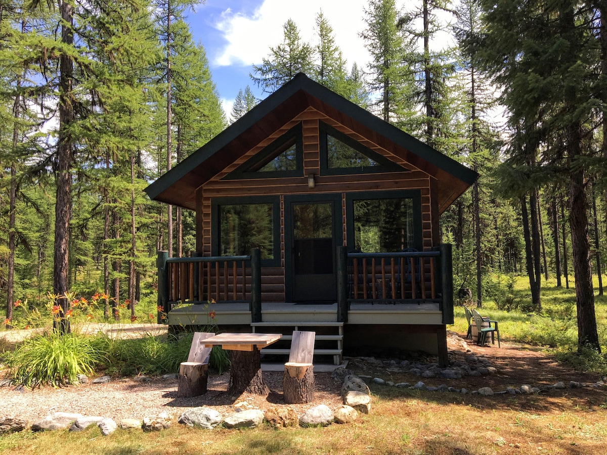 A cozy cabin nestled among tall trees demonstrates a rustic charm. The deck features seating for relaxation, while a stone pathway leads to a simple wooden table for outdoor dining. Surrounding greenery complements the serene atmosphere of the secluded setting.