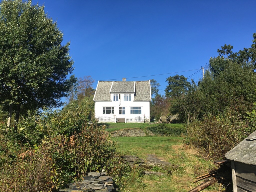 The exterior of a charming white house with a sloped roof is framed by green foliage and trees. A clear blue sky provides a bright backdrop, highlighting the tranquil setting. A well-maintained lawn leads up to the house, inviting guests to explore.