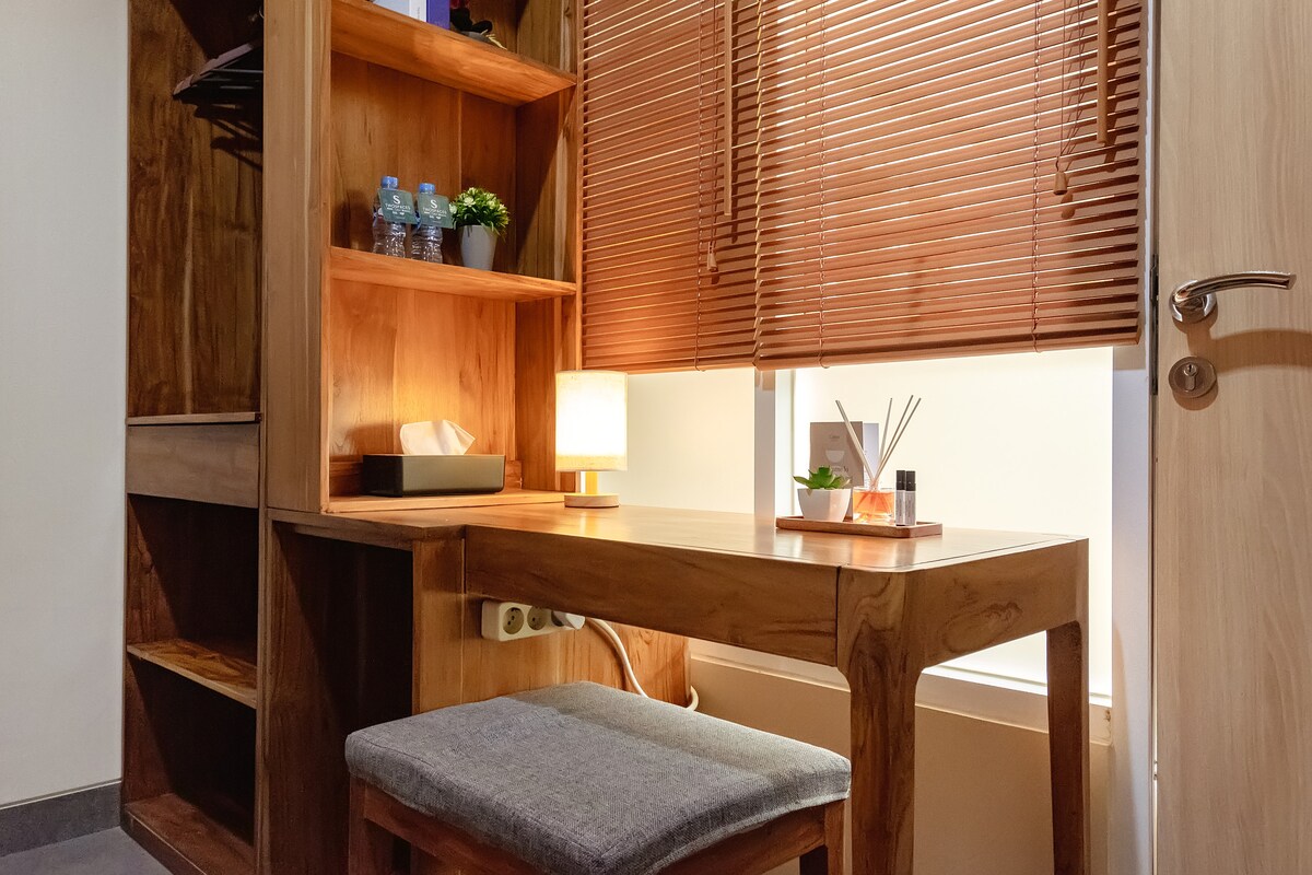 A wooden desk is positioned next to a window, featuring a small lamp and decorative items. A chair with a cushioned seat complements the desk. Natural light filters through wooden blinds, creating a warm, inviting space. Shelves hold plants and bottled water, enhancing functionality.