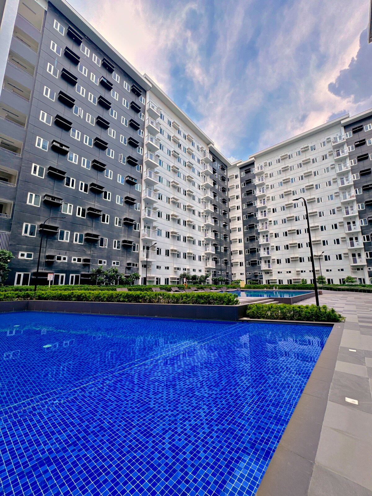 A view of the resort-style swimming pool is captured, featuring crystal-clear water surrounded by lush greenery. Modern condominium buildings rise in the background, displaying a blend of dark and light exterior finishes. The sky is partly cloudy, adding to the serene atmosphere.