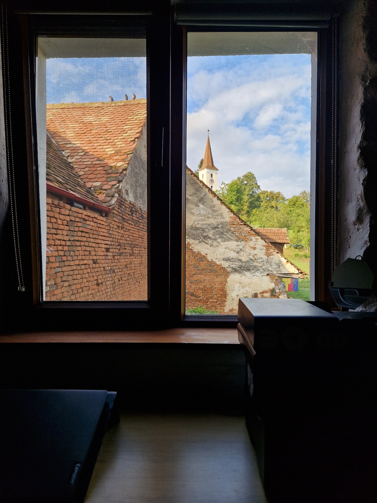 A view from the window reveals a charming rural landscape featuring a historic building's textured brick exterior and a church spire reaching toward the sky. Lush greenery frames the scene, with soft clouds scattered across the blue backdrop.