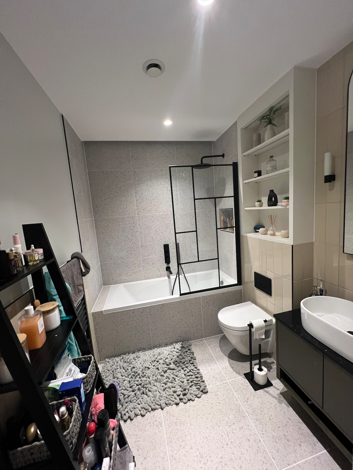 The bathroom features a modern design with a glass shower enclosure and a spacious bathtub integrated into the tiled wall. A vanity with a round sink is placed beside a sleek toilet. Shelves display various toiletries and decorative items, while a textured rug adds warmth to the floor.